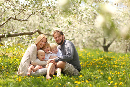 A portrait of a happy family of five people, including mother, father, 2 young boy children and baby girl holding hands and hugging outside in a flower meadow under blossoming apple trees on a spring day.